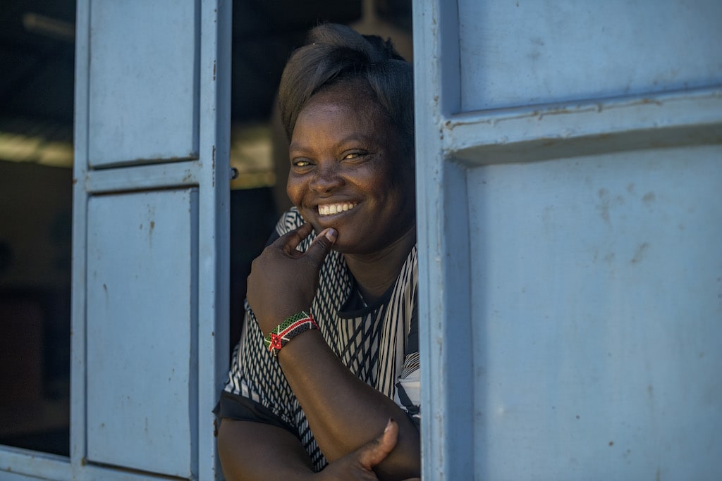 Florence is wearing a black and white dress. She is looking out of a window with blue shutters at the Compassion centre.